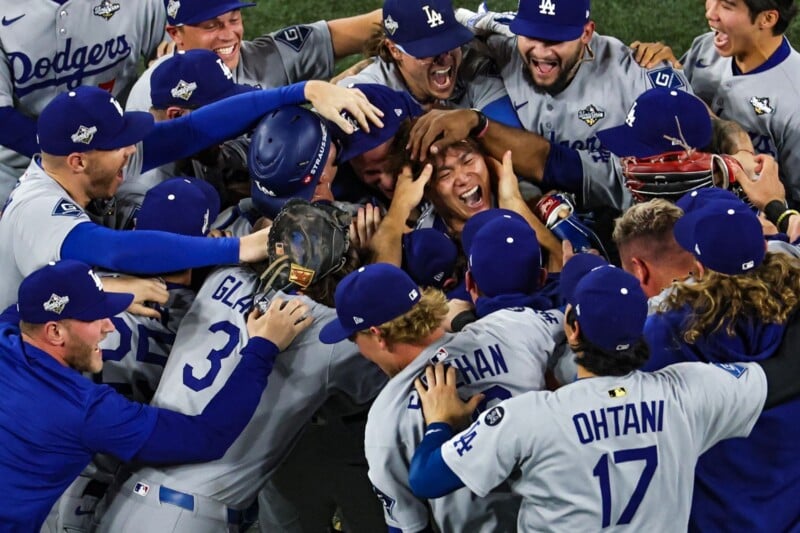A group of Los Angeles Dodgers baseball players in grey uniforms joyfully celebrate on the field, hugging and smiling in a close, energetic huddle after a victory.