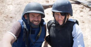 Two people wearing blue body armor and black helmets stand close together on a sandy area. One person has an eye patch. They both look towards the camera and appear to be in a conflict or war zone.