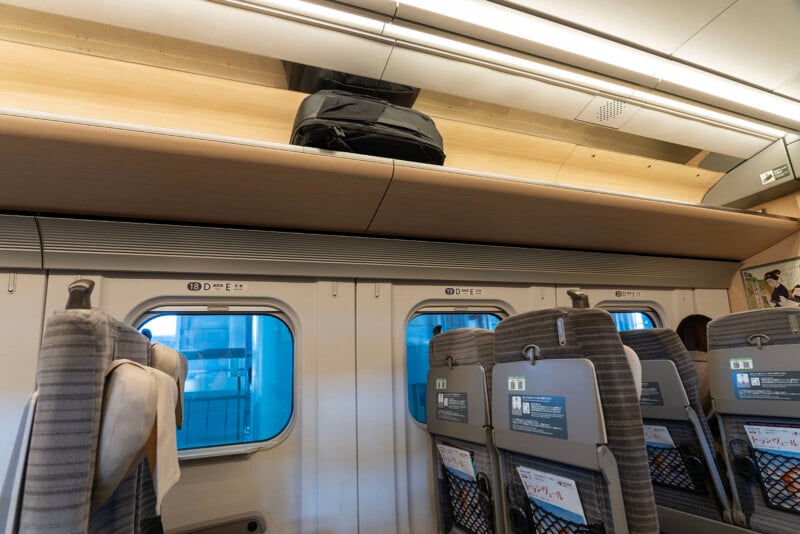 A black bag sits on the overhead luggage shelf inside a modern train, above gray passenger seats and large windows with blue reflections.