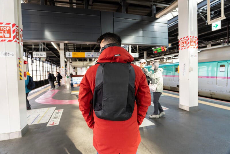 A person wearing a red jacket and black backpack stands on a train platform, facing away. Other people and a train are visible in the background under bright lighting. Signs and directions are on the walls and floor.