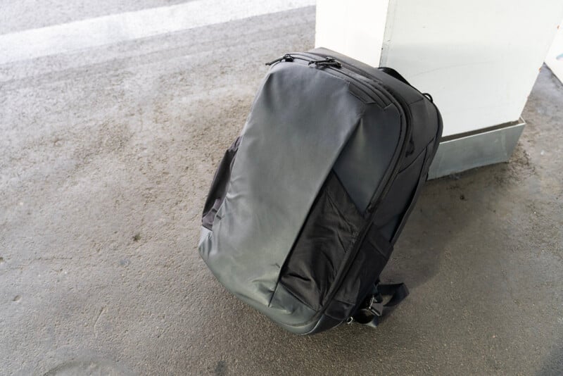 A large black travel backpack rests upright against a white pillar on a textured concrete surface, possibly at a transit station or parking area.