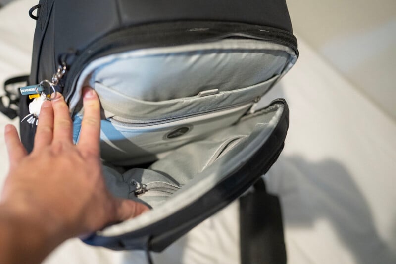 A hand holds open a black backpack, revealing the light gray interior with several zippered compartments and pockets, displayed on a white surface.