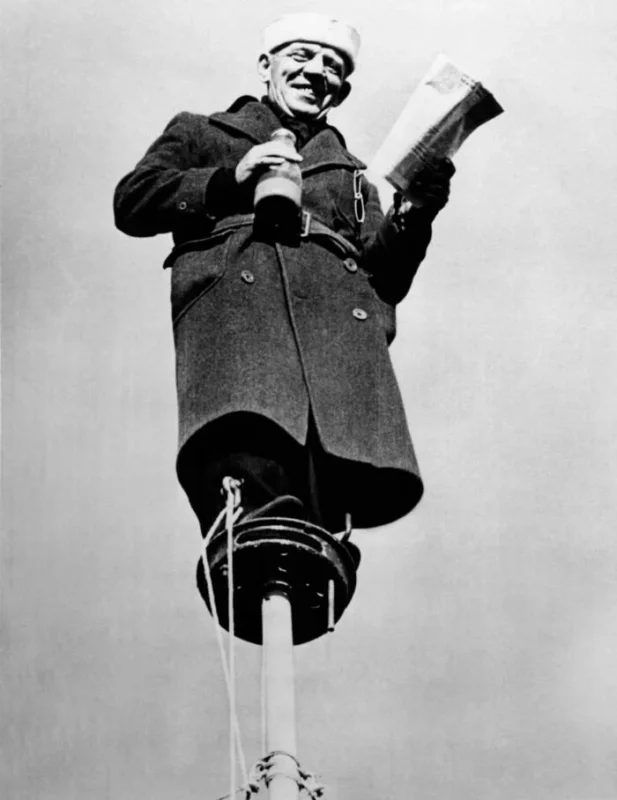 A sailor in a long coat and hat stands on top of a ship’s mast, holding a paper and a bottle, looking down, with a clear sky in the background. The image is taken from a low angle.