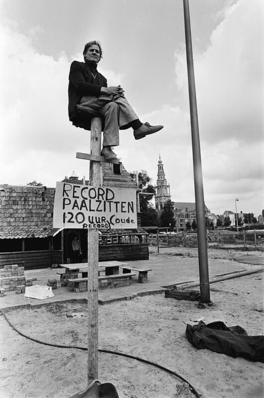 A man sits on top of a tall wooden pole, legs crossed, above a sign that reads "Record Paalzitten 120 uur Oude record" in Dutch. Buildings and a church tower are visible in the background.