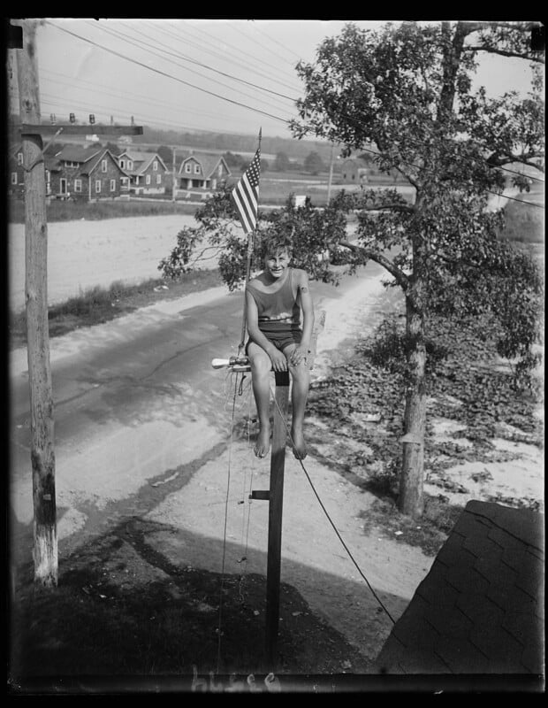 A young person in a swimsuit sits on top of a tall pole with an American flag, above a rural road, with trees and houses in the background on a sunny day.