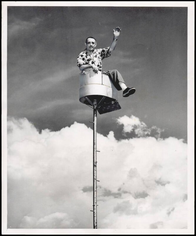 A man sits in a small cylindrical seat atop a tall pole, waving with one hand against a backdrop of clouds and blue sky. He appears relaxed and is dressed in casual clothing and sunglasses.