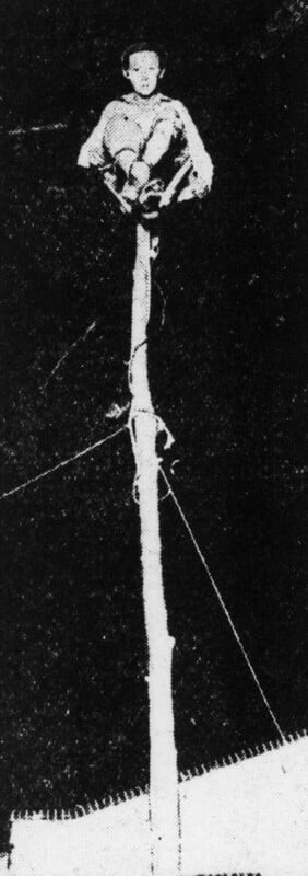 A person sits at the top of a tall, narrow pole at night, with their knees drawn up and arms resting on their knees. The surroundings are dark except for the pole and the seated figure.