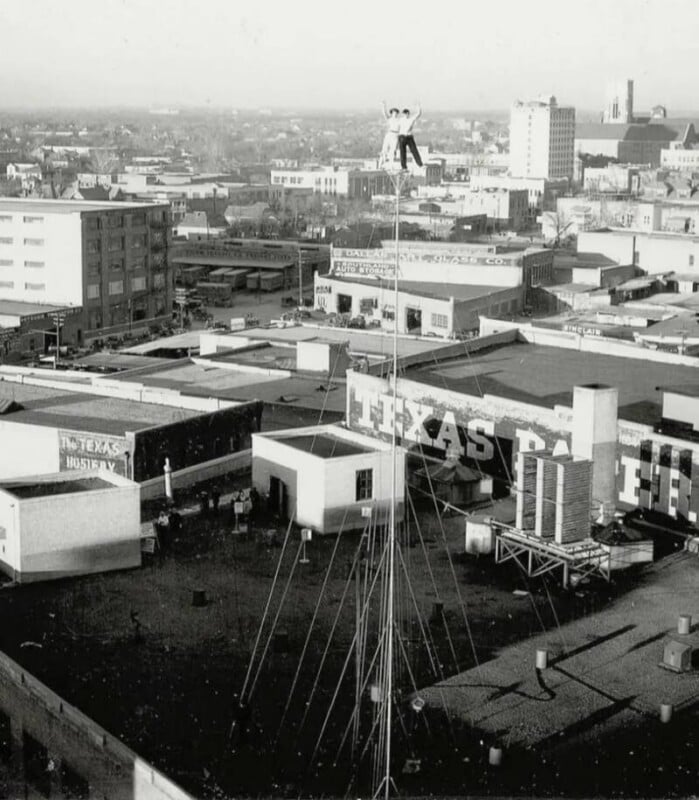 A person stands high atop a thin pole above a cityscape, surrounded by buildings and rooftops, with large signs reading "TEXAS" visible on nearby structures.
