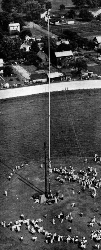 Aerial view of a large crowd gathered around a tall pole being erected in an open grassy field, with houses and roads visible in the background.
