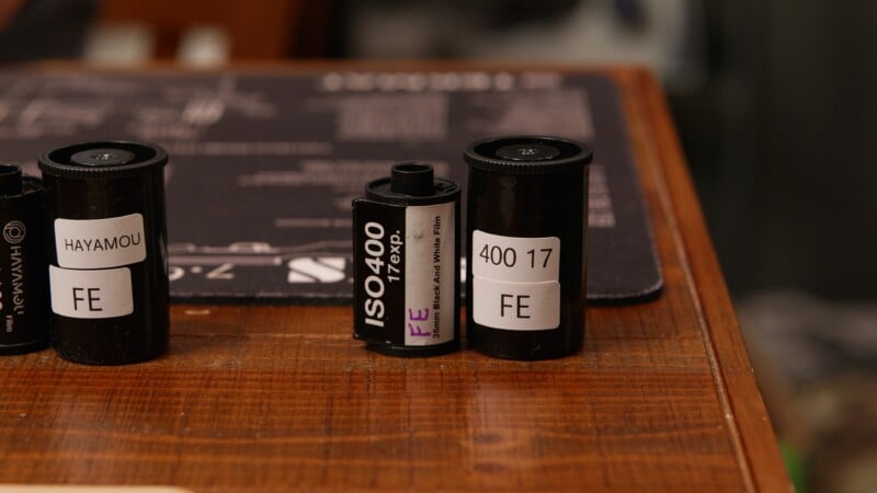 Three film canisters and one exposed 35mm film roll labeled with "HAYAMOU," "ISO400 79XP.," and "400 17 FE" sit on a wooden surface with a dark mat in the background.