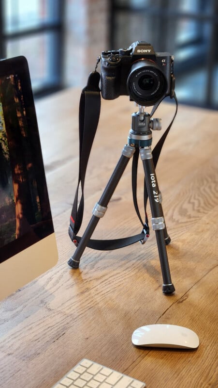 A Sony camera on a tripod is set up on a wooden desk next to a computer monitor, wireless keyboard, and mouse in a bright, modern office setting.