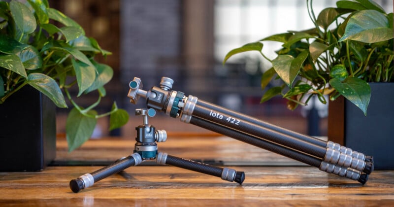 A carbon fiber tripod labeled "iOta 422" stands partially extended on a wooden table between two potted green plants, with a blurred indoor background.