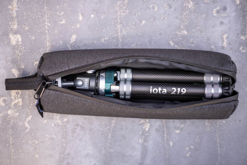 A black cylindrical device labeled "iota 210" is packed inside a partially unzipped, dark gray fabric case lying on a textured gray surface.