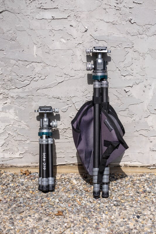 Two black and silver tripods stand upright against a textured gray wall; one is folded and compact, the other is extended and has a dark carrying case attached. The tripods are on a pebbled ground.