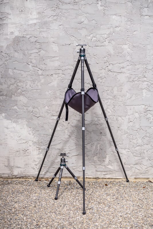 Two black tripods stand on a gravel surface against a textured gray wall; the larger tripod holds a dark bag between its legs, while the smaller tripod is positioned in front of it.