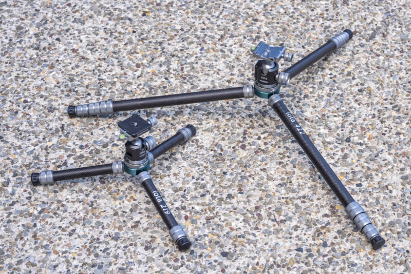 Two black and silver tripod bases with spread legs rest on a speckled, textured concrete surface. Each tripod has a mounting plate on top and appears to be made of carbon fiber.