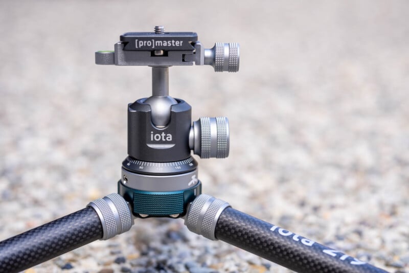 A close-up of a ProMaster iota tripod head with metallic adjustment knobs, attached to carbon fiber tripod legs, set on a textured, gravel surface.