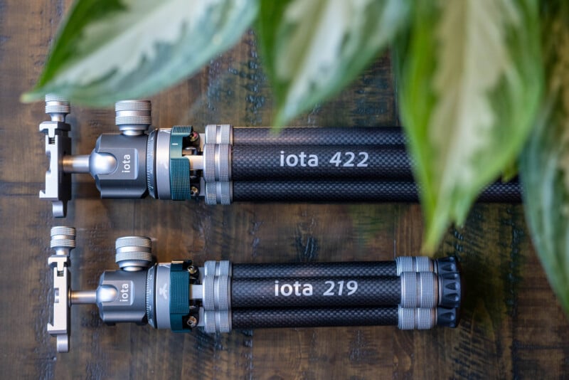 Two black tripods labeled "iota 422" and "iota 219" lie side by side on a wooden surface, partially covered by green leaves in the upper right of the image.
