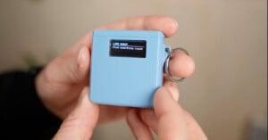 A person holds a small blue electronic device with a keyring. Its screen displays the text: "LIFE QUEST Find something round.