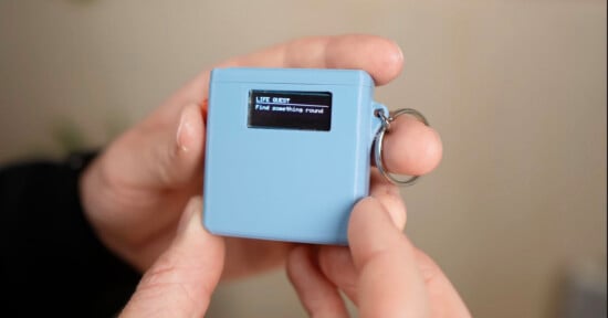 A person holds a small blue electronic device with a keyring. Its screen displays the text: "LIFE QUEST Find something round.