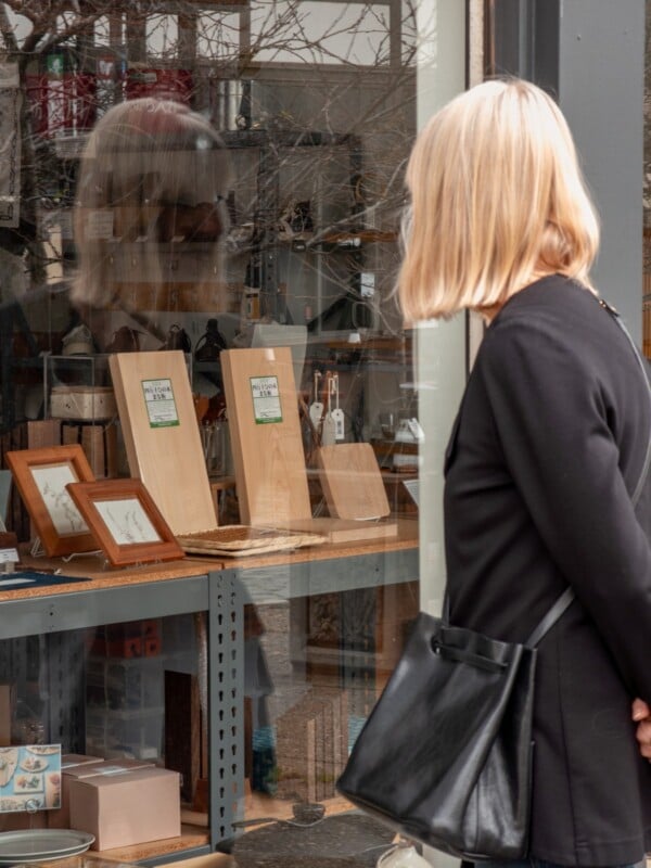 A woman with blonde hair, wearing a black outfit and carrying a black purse, looks into a shop window displaying wooden cutting boards, framed pictures, and various small items.