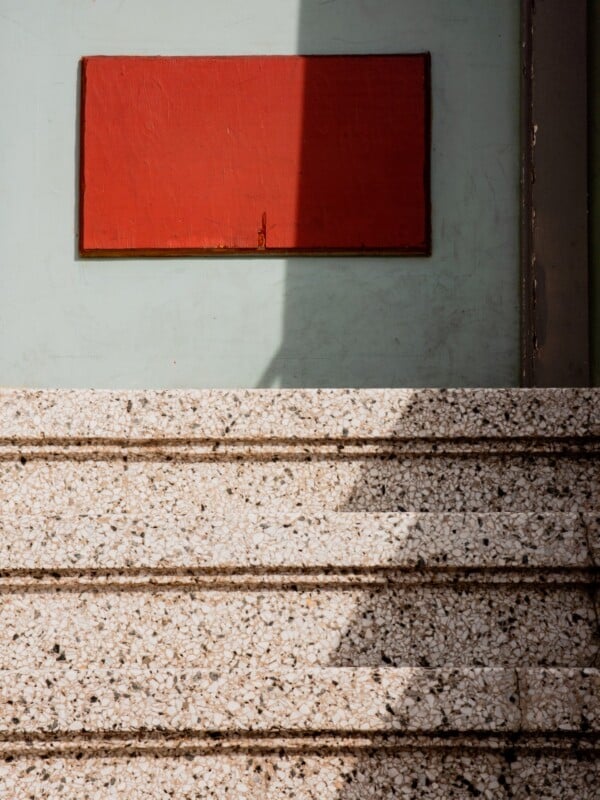 A textured stone staircase leads up to a pale green wall with a large rectangular red panel; sunlight and shadows create diagonal lines across the scene.