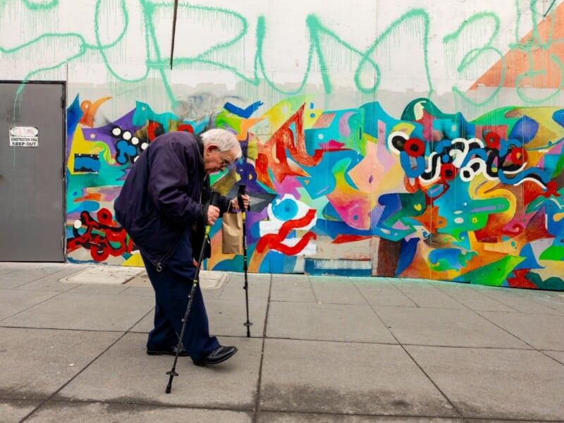 An elderly man using two walking sticks passes by a colorful, abstract graffiti mural on a city sidewalk. He is carrying a brown paper bag and appears to be focused on his steps.