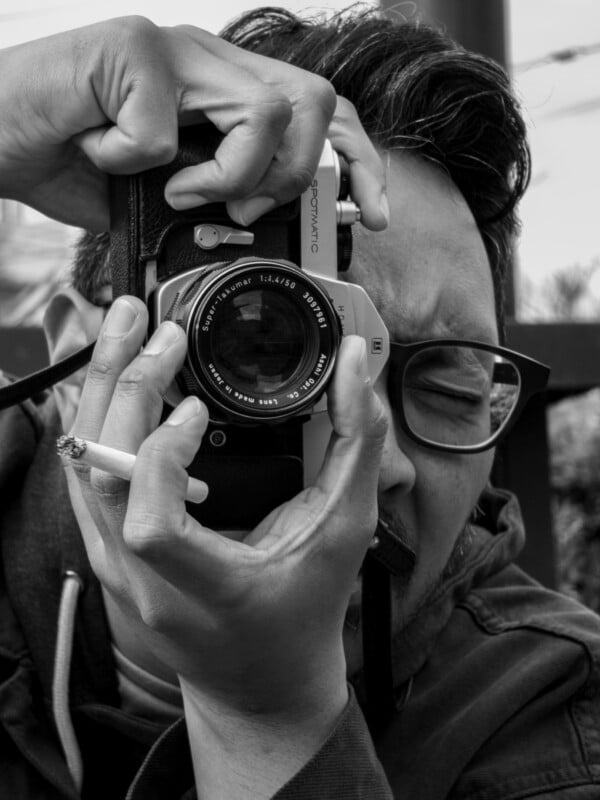 A person wearing glasses holds a vintage camera up to their eye, preparing to take a photo. They have a cigarette between their fingers. The image is in black and white.