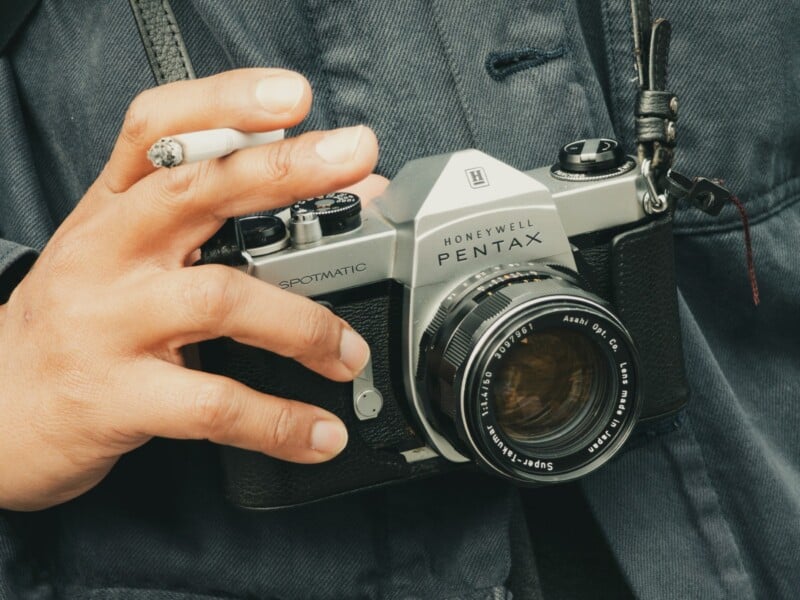 A person holding a vintage Honeywell Pentax Spotmatic film camera with one hand while holding a lit cigarette between their fingers, wearing a dark jacket.