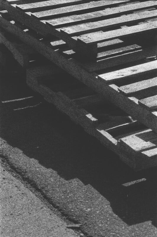 Black and white photo of wooden pallets stacked on top of each other, casting shadows on the ground. The image captures the texture of the wood and rough surface of the pavement below.