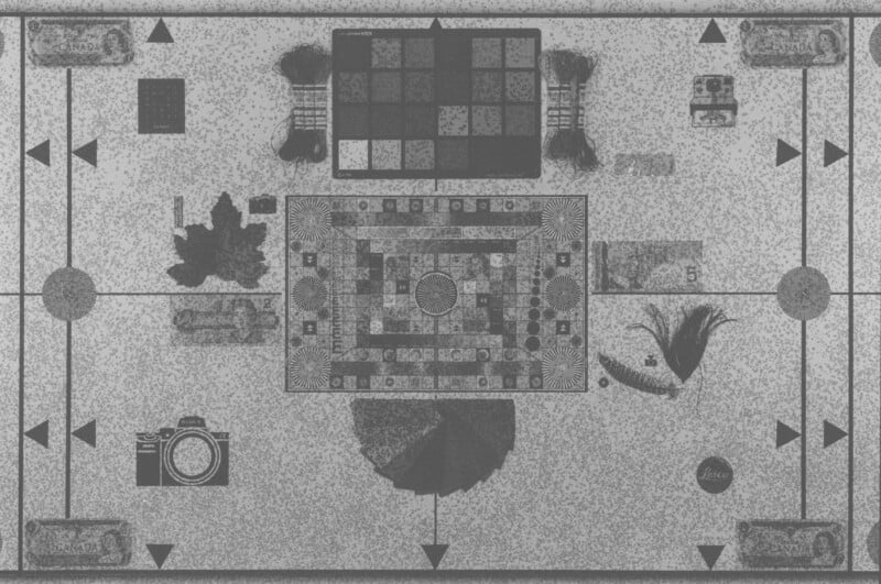 A grayscale image of a patterned board game with geometric shapes, squares, arrows, and various illustrations including cameras, a leaf, currency notes, and feathers arranged around the central board.