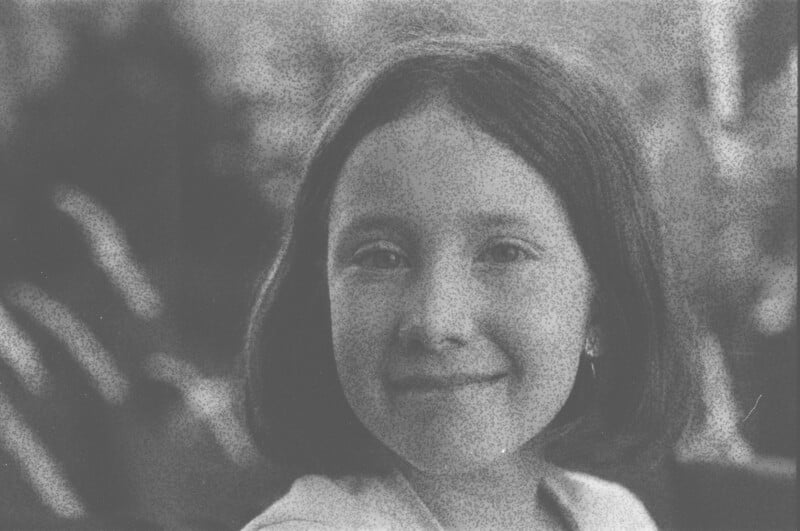 A young girl with short dark hair smiles softly at the camera. The image is in black and white with a grainy texture, and the background is blurred.