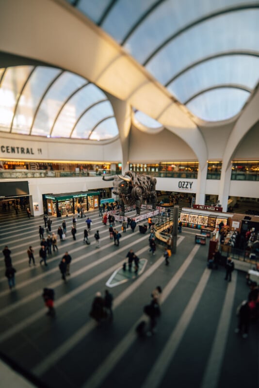 Wide interior view of a modern train station with a high, curved glass ceiling. People walk and gather below, with shops and a large bull statue visible in the central area.