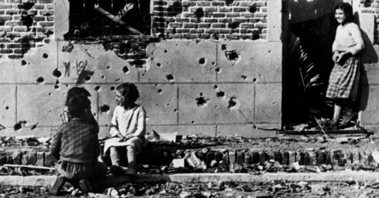 Three children sit on a curb in front of a building wall riddled with bullet holes; another child stands in a doorway, all appearing somber. Debris and rubble are scattered on the ground.