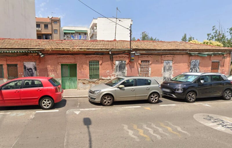 Three cars—a red, a silver, and a black—are parked on a street in front of a long, old brick building with green doors and windows. The building appears worn, with faded posters and peeling paint.