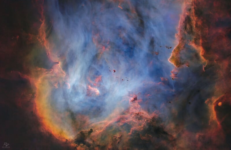 Una nebulosa colorida con nubes arremolinadas de gas azul, naranja y rosa; contornos oscuros de gas y polvo parecen formar el contorno de una cara en el lado derecho de la imagen.