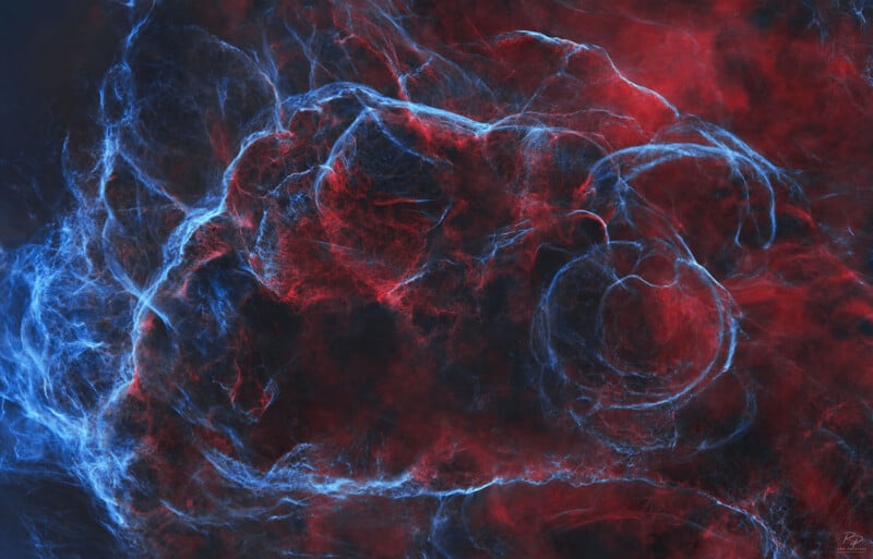 Nubes arremolinadas de luz roja y azul se entrelazan contra un fondo oscuro, creando un patrón abstracto etéreo y nebular con texturas delicadas y tenues.