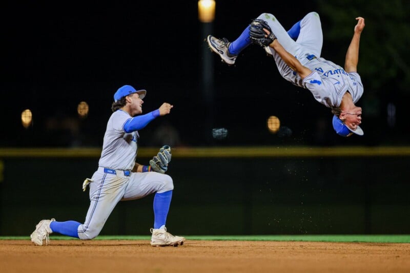Two baseball players in blue and white uniforms celebrate on a field at night; one is kneeling and cheering, while the other is mid-air performing a backflip, both appearing joyful.