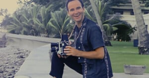 A smiling man in a short-sleeve button-up shirt kneels outdoors by the ocean, holding a vintage movie camera, with palm trees and a building in the background.