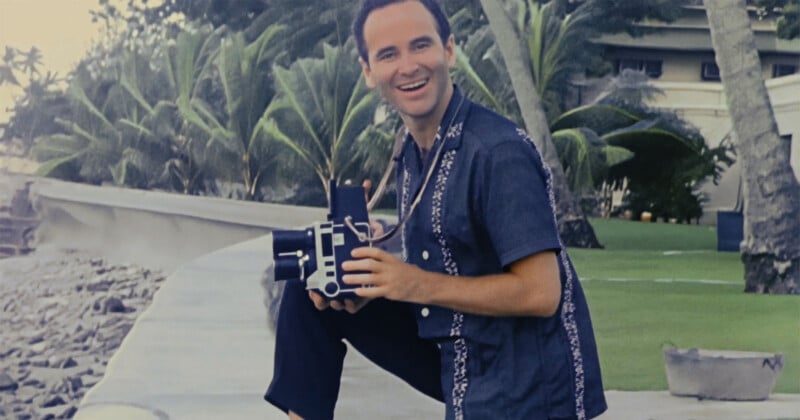 Un hombre sonriente con una camisa de manga corta abotonada se arrodilla al aire libre junto al mar y sostiene una cámara de cine antigua con palmeras y un edificio al fondo.