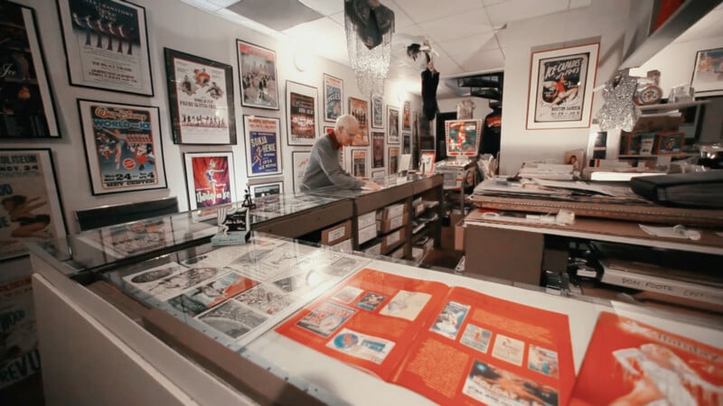 Una persona se encuentra detrás del mostrador de una tienda de souvenirs o de un archivo, rodeada de carteles, fotografías enmarcadas y vitrinas llenas de objetos y documentos antiguos. El espacio parece organizado pero ocupado.