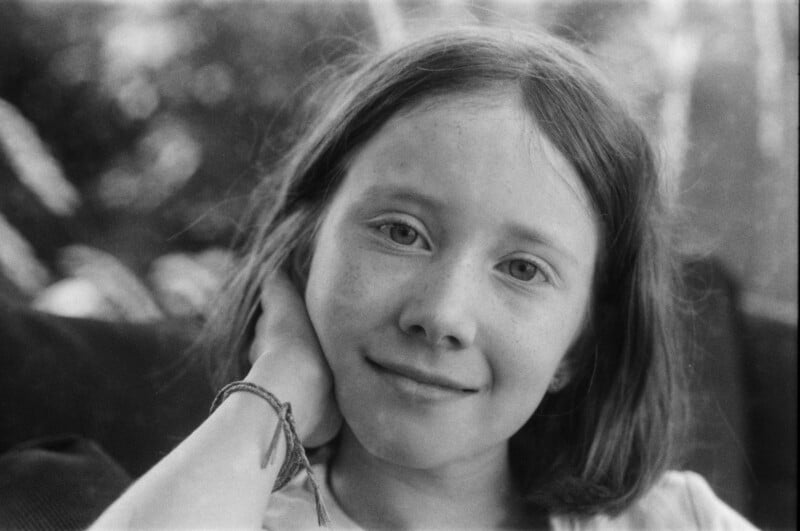 A young girl with light skin and straight hair smiles softly at the camera, resting her head on her hand. She is wearing a bracelet, and the background is blurred with natural light.