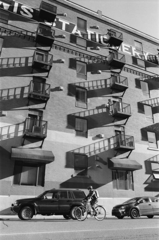 A person rides a bicycle past parked cars on a city street, alongside a brick building with multiple small balconies casting long shadows in the sunlight.