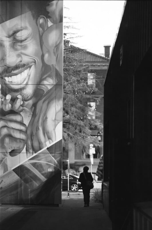 A person walks down an alley next to a building featuring a large mural of a smiling face. Trees, cars, and a brick building are visible in the background. The image is in black and white.