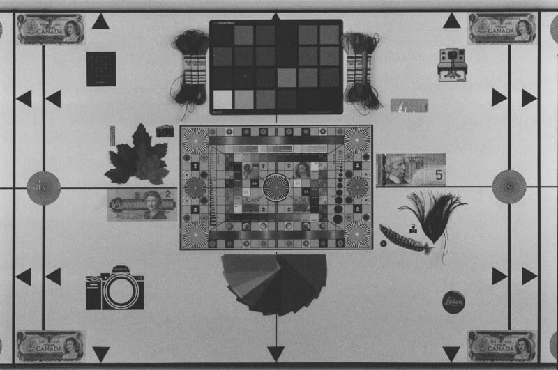 A black and white image displays various objects on a flat surface, including Canadian currency, maple leaves, a camera illustration, film rolls, a feather, color charts, and measuring scales arranged symmetrically.