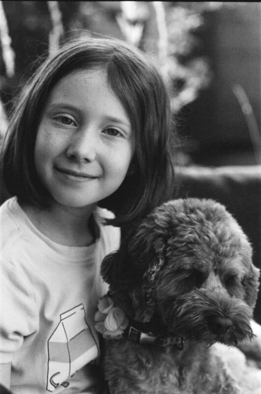 A young child with shoulder-length hair smiles at the camera while holding a curly-haired dog. The photo is black and white, and the background is softly blurred.