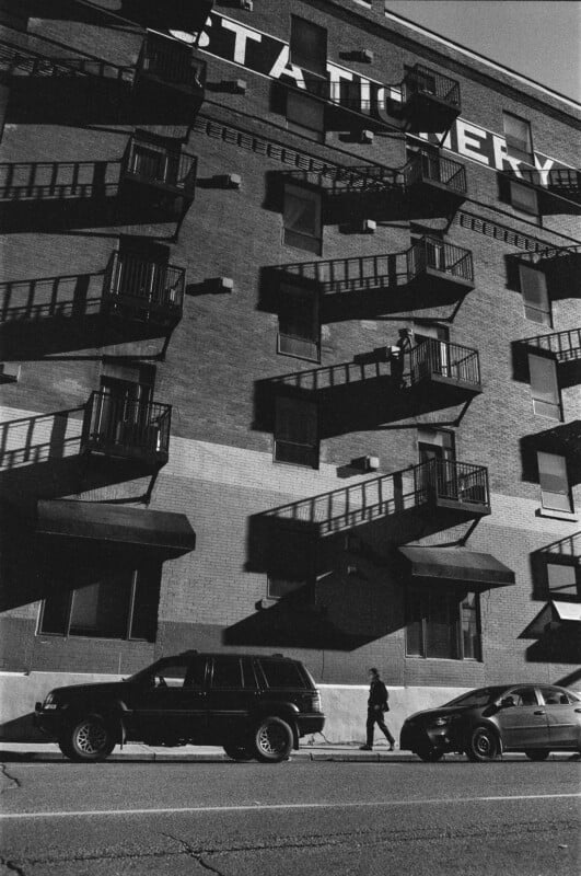 A black and white photo shows a person walking on the sidewalk beside two parked cars in front of a brick building with fire escape shadows and a large, partially visible “STATIONERY” sign at the top.
