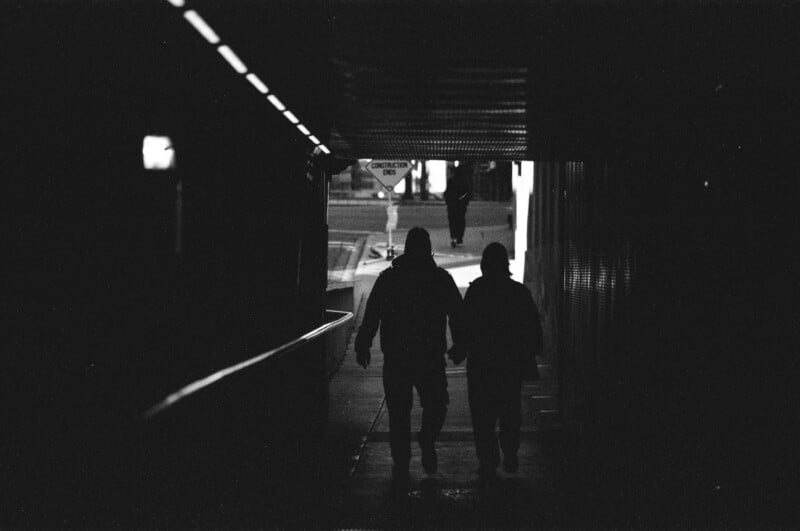 Two people walk hand in hand through a dark tunnel toward a brighter street outside, with another person visible in the distance. The scene is in black and white, with strong contrast between shadows and light.