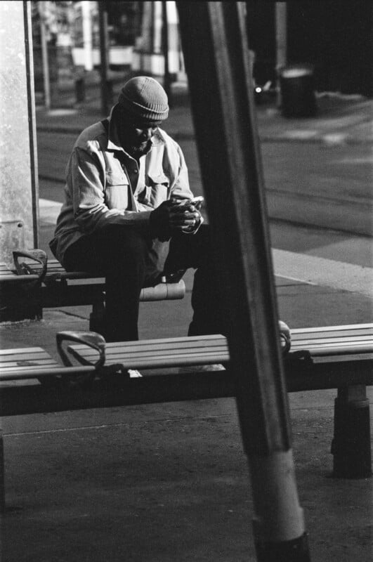 A man wearing a beanie and sunglasses sits alone on a bench at a transit stop, looking down at his phone, in a black-and-white urban setting.
