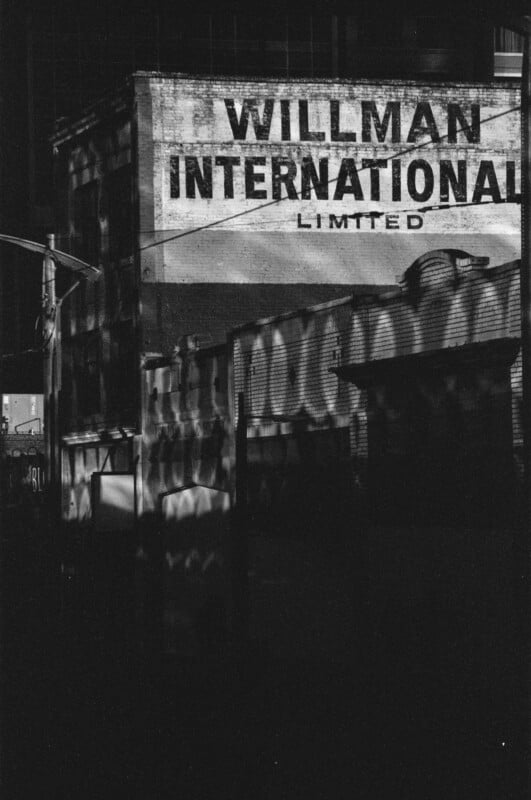 A black and white photo of an old building with a faded sign reading "WILLMAN INTERNATIONAL LIMITED." Shadows and light patterns are cast across the brick walls, creating an atmospheric, moody scene.